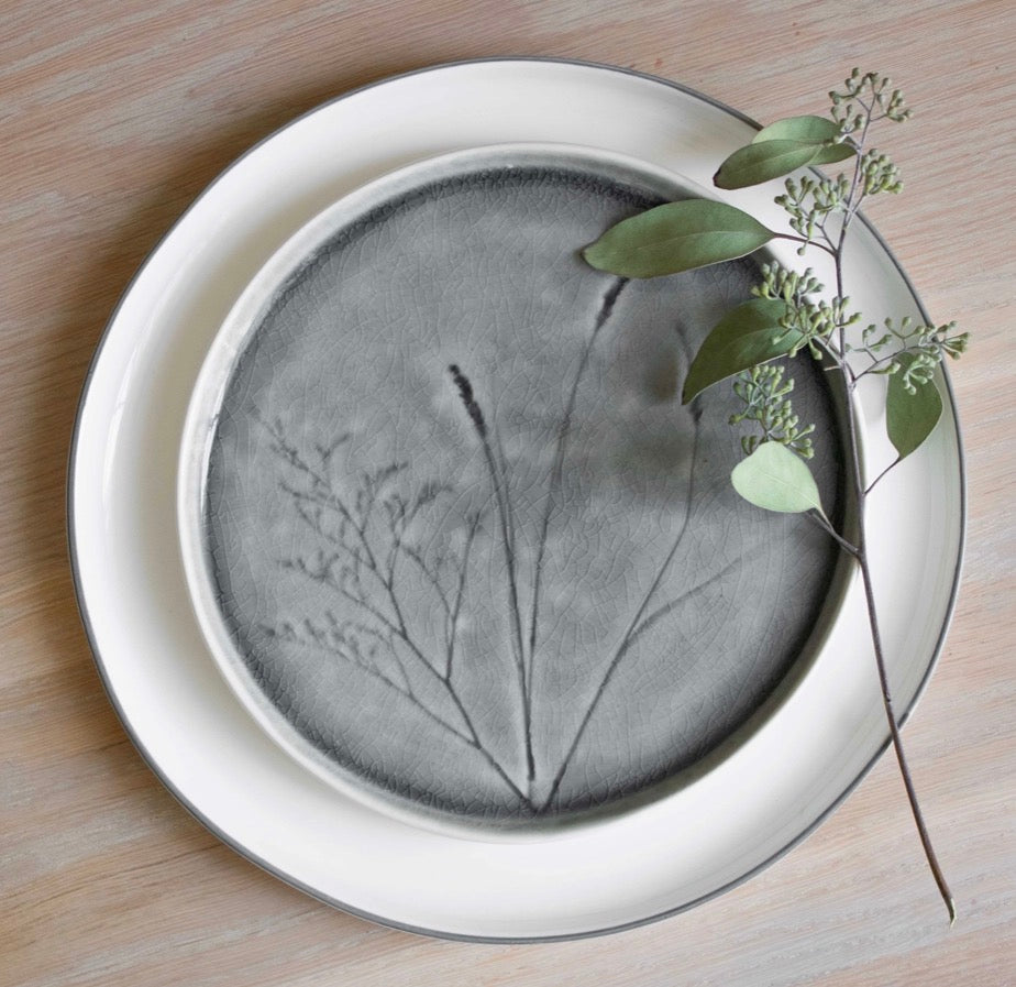 A round ceramic plate with a dark gray botanical leaf print, layered on top of a larger white charger plate with a thin gray rim, styled with a green eucalyptus sprig on a light wood background.