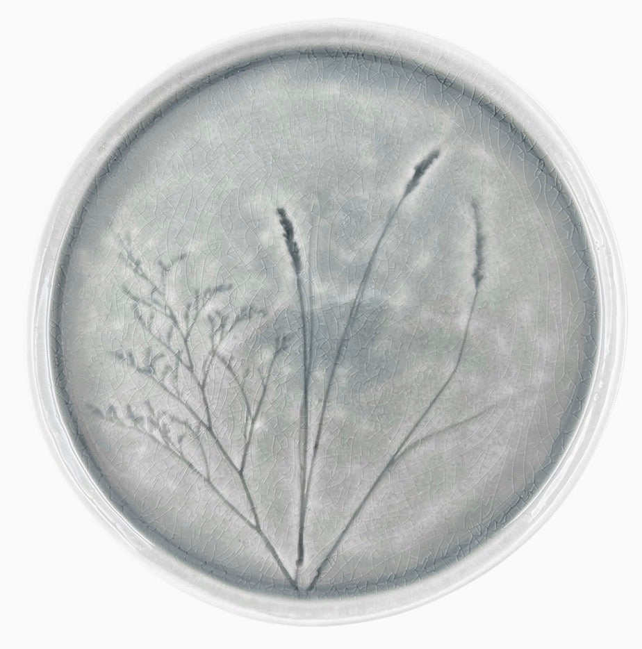 A circular gray art print or plaque featuring a botanical design of three thin grass-like stems on a textured gray background, surrounded by a clear circular border.