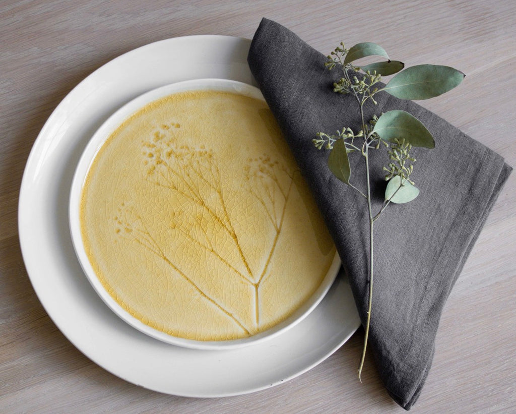 A top-down view of a yellow ceramic plate with a botanical branch pattern, nested inside a larger white plate, styled with a dark gray linen napkin and a green eucalyptus sprig on a light wood tabletop.