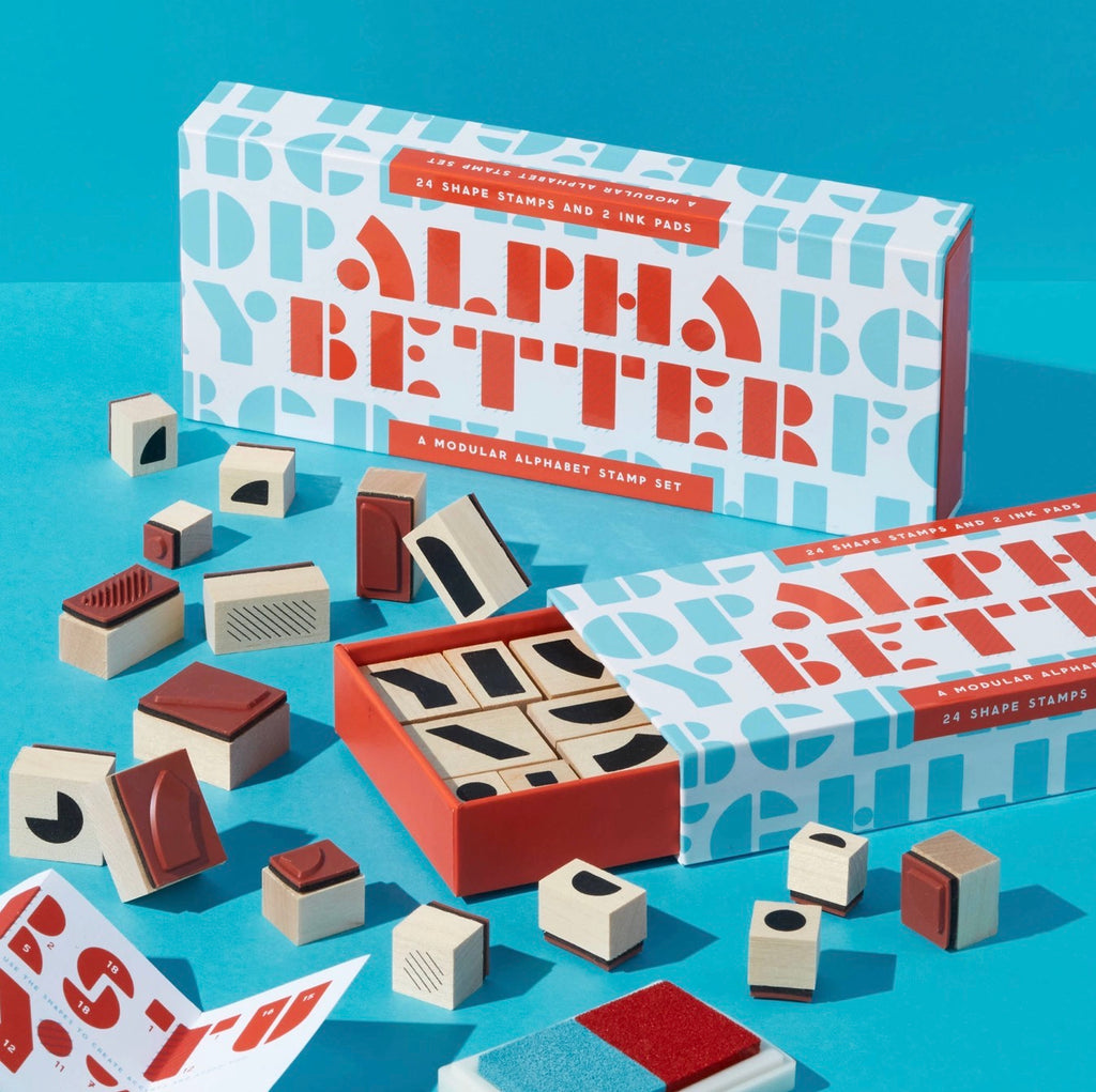 Alpha Better alphabet stamp set with two patterned boxes, an open box showing wooden stamps, and several loose stamp blocks arranged on a bright blue background.