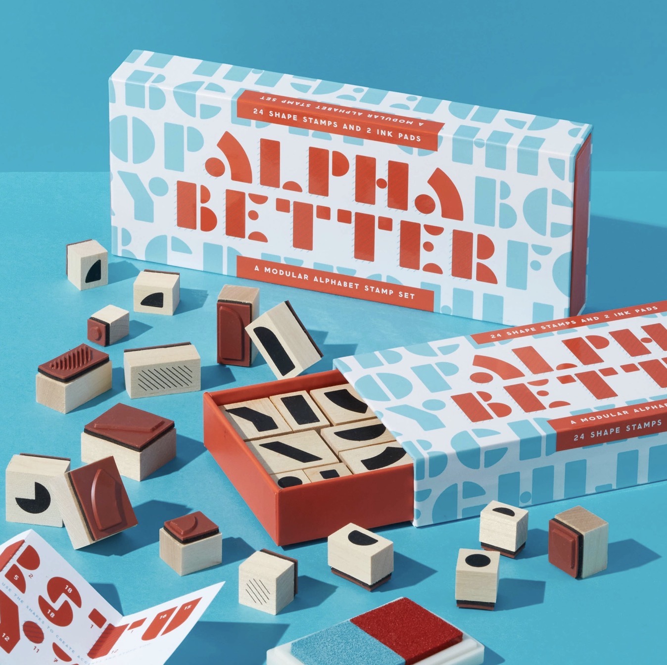 Alpha Better alphabet stamp set with two patterned boxes, an open box showing wooden stamps, and several loose stamp blocks arranged on a bright blue background.