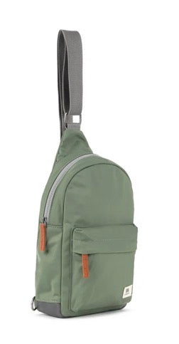 An olive green sling backpack with a gray adjustable shoulder strap, gray zipper trim, and small brown leather accents on the zipper pulls and a small white brand label on the lower right front pocket.