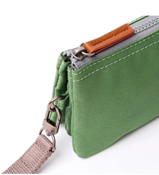 A close-up of a green canvas zip pouch with a brown leather zipper pull, silver metal hardware, and a matching green wrist strap on a white background.