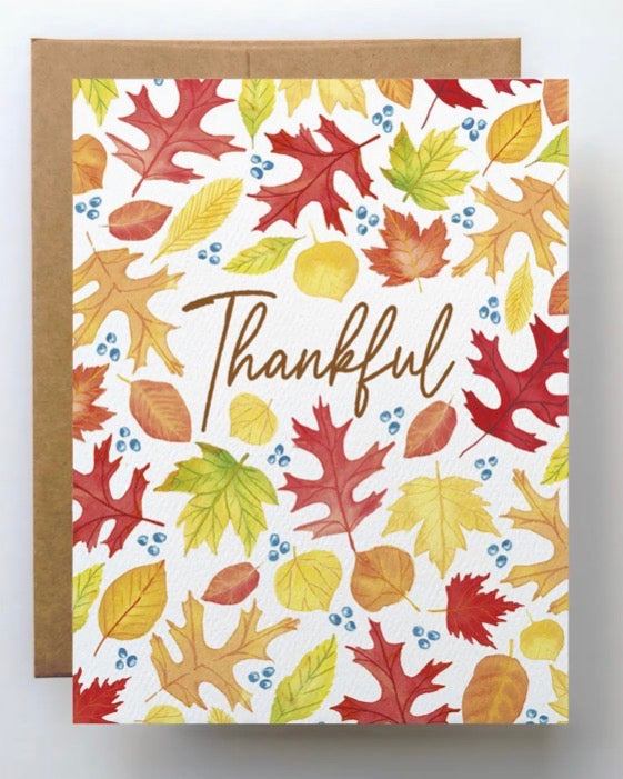 A square greeting card with a white background covered in colorful autumn leaves and small blue berries, with the word 'Thankful' written in brown script across the center. A kraft brown envelope is placed behind the card on a light gray background.