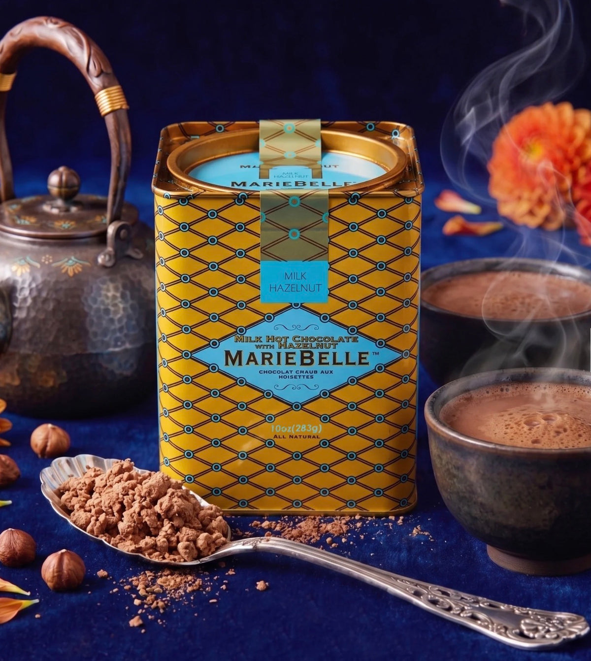 A tin of MarieBelle Milk Chocolate Cacao Confectionery sits on a dark blue surface with a spoon of chocolate powder, whole chocolate hazelnuts, two cups of hot chocolate with steam, and a metal teapot in the background.