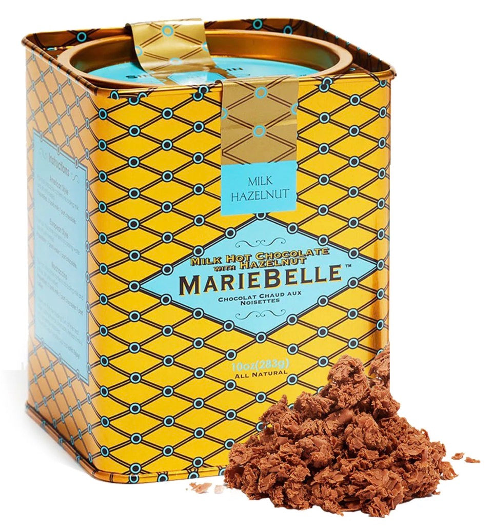 A vintage-style MarieBelle milk hazelnut chocolate tin with a yellow and blue geometric pattern, shown with a pile of chocolate hazelnut cookie crumbs in front on a white background.
