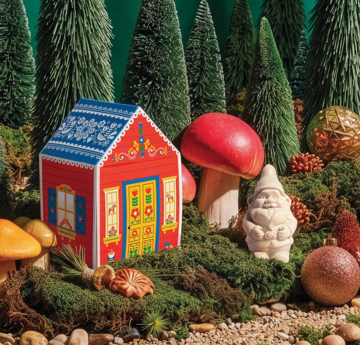 A decorative red gingerbread-style house ornament displayed in a miniature forest scene with artificial pine trees, a large red mushroom, a small gnome statue, moss, pinecones, and colorful ornaments.