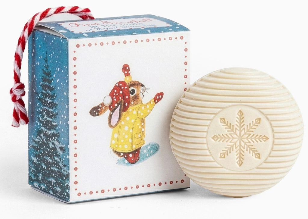 A round cream-colored textured soap bar with an embossed gold snowflake, displayed next to its illustrated Christmas-themed gift box featuring a rabbit in a yellow coat.
