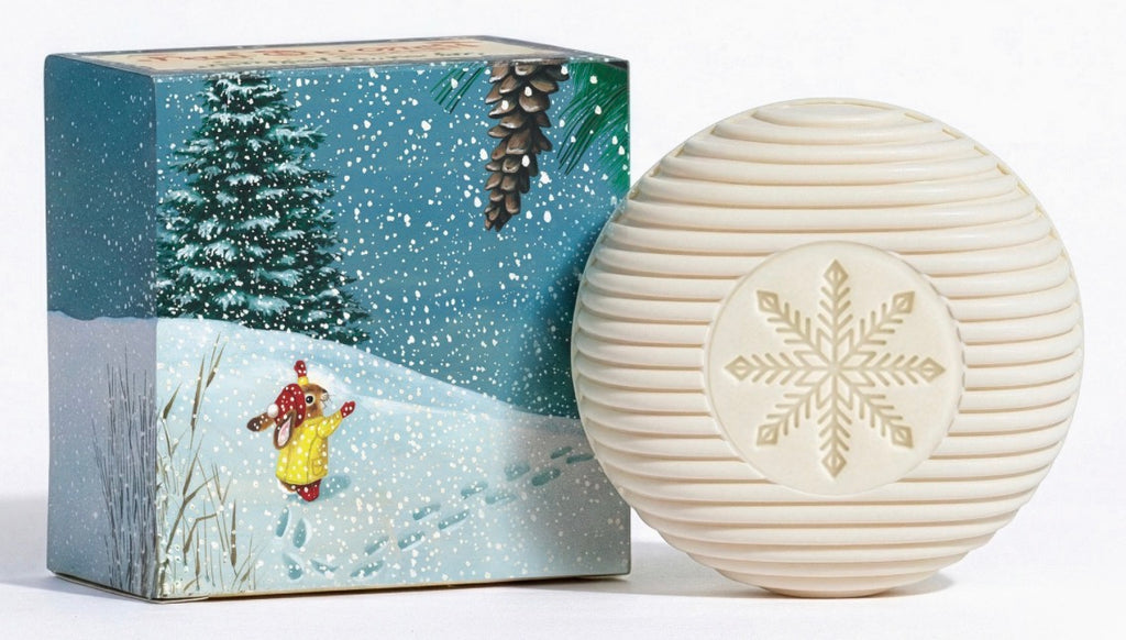 A round cream-colored ribbed soap or shampoo bar with a gold snowflake design, shown next to its illustrated winter-themed box featuring a snowy landscape and a child in a yellow coat.