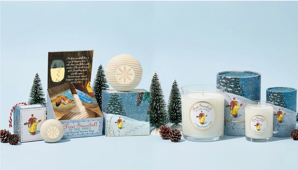 A coordinated set of winter-themed candles and gift boxes arranged on a pale blue studio background. The products include a large white candle in a clear glass jar, a smaller matching candle, cylindrical blue patterned candle holders, a round cream candle, and wrapped gift boxes with printed labels and small decorative evergreen trees and pinecones.