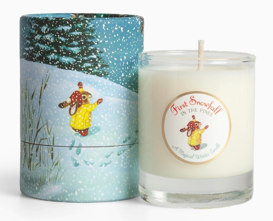 A scented candle in a clear glass jar next to its illustrated cylindrical tin. The candle wax is off-white, and the label reads 'First Snowfall in the Pines'.