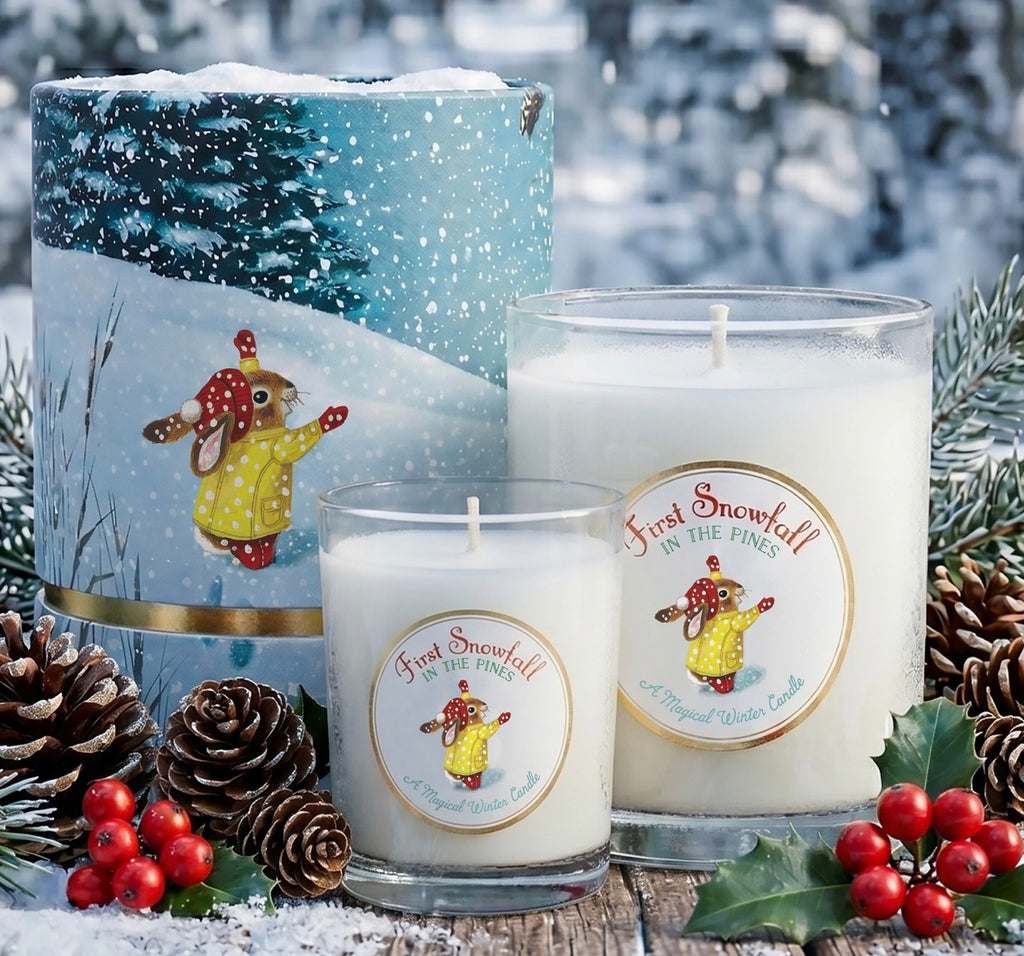 Three First Snowfall candles displayed in a snowy outdoor-style setting: a large cylindrical tin, a medium glass jar candle, and a small glass candle. The candles are decorated with round labels featuring a rabbit in a yellow coat, surrounded by pinecones, red berries, and holly.