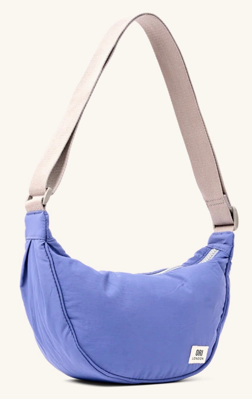 A blue crescent-shaped shoulder bag with a beige adjustable strap, shown against a plain off-white background. A small white brand tag is visible on the lower right corner of the bag.
