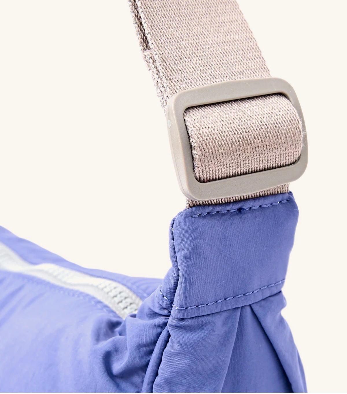 Close-up of a blue sling bag strap with a gray plastic buckle and a light beige woven strap, set against a plain off-white background.