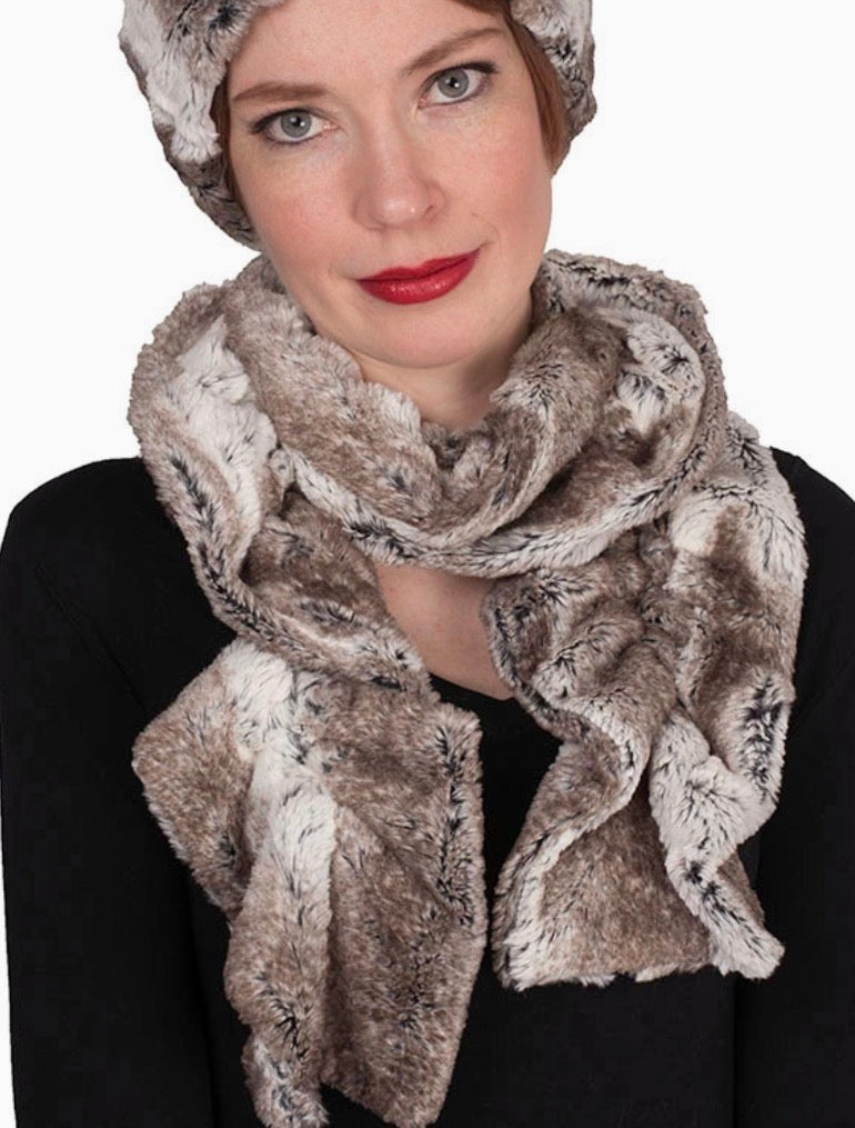 A woman wearing a plush faux-fur style scarf and matching beanie in taupe, brown, and cream tones, paired with a black top against a plain white background.