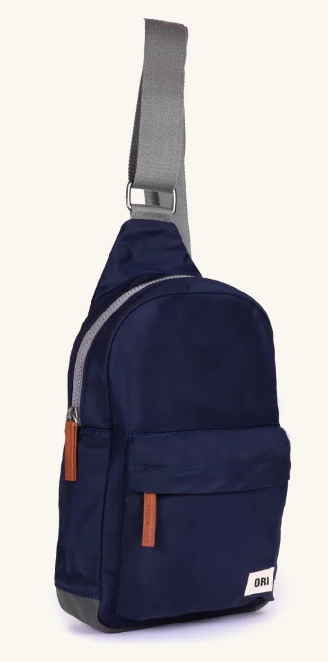 A navy blue sling backpack with a gray adjustable shoulder strap, brown leather accents, and a small white 'Ora' brand patch on the front pocket, set against a plain off-white background.