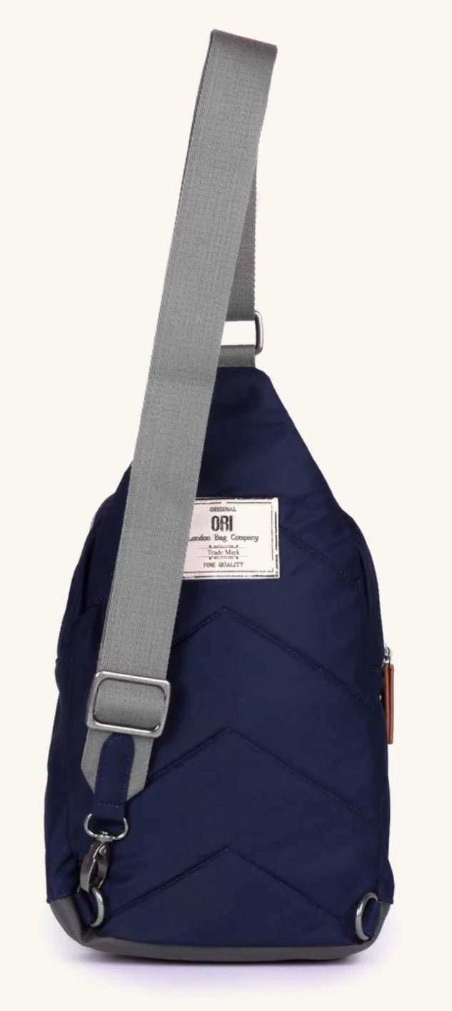 A navy blue sling backpack with a gray adjustable shoulder strap and a white brand label on the front, shown against a plain off-white background.