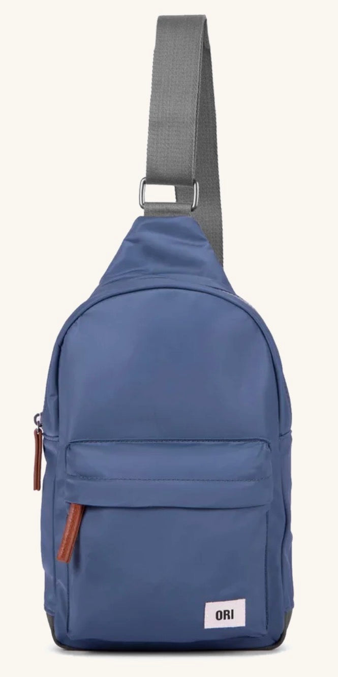 A blue sling backpack with a gray adjustable shoulder strap, featuring a front zippered pocket with brown leather zipper pulls and a small white ORI brand label on the bottom right.