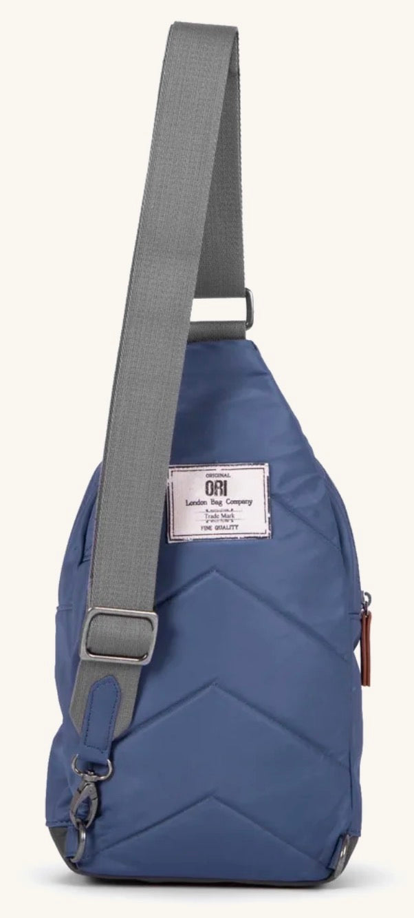 A blue sling backpack with a gray adjustable shoulder strap and a white brand label on the front, shown against a light off-white background.