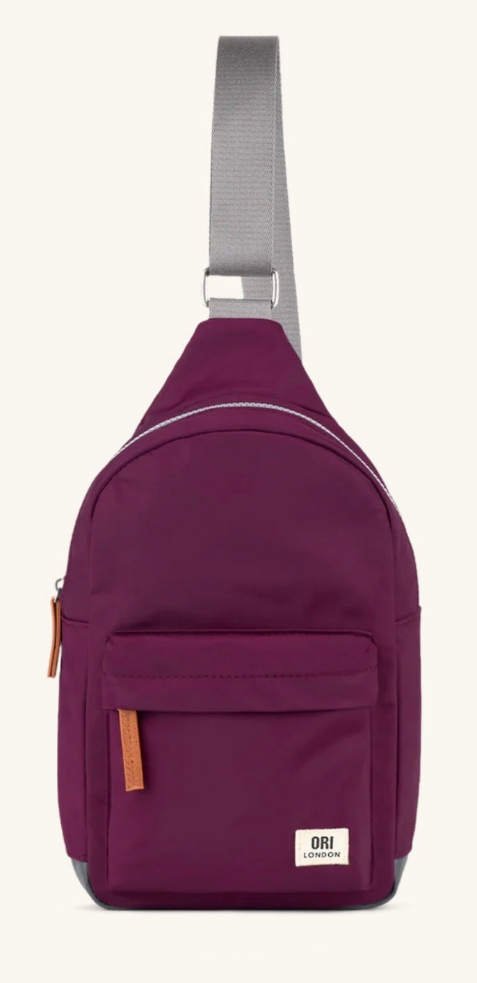 A burgundy sling backpack by Ori London with a gray adjustable shoulder strap, front zippered pocket, and a small white brand label on the bottom right.