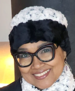 A close-up portrait of a smiling woman wearing a chunky black and white knit hat with a fluffy black band, black cat-eye glasses, and a white knit scarf.