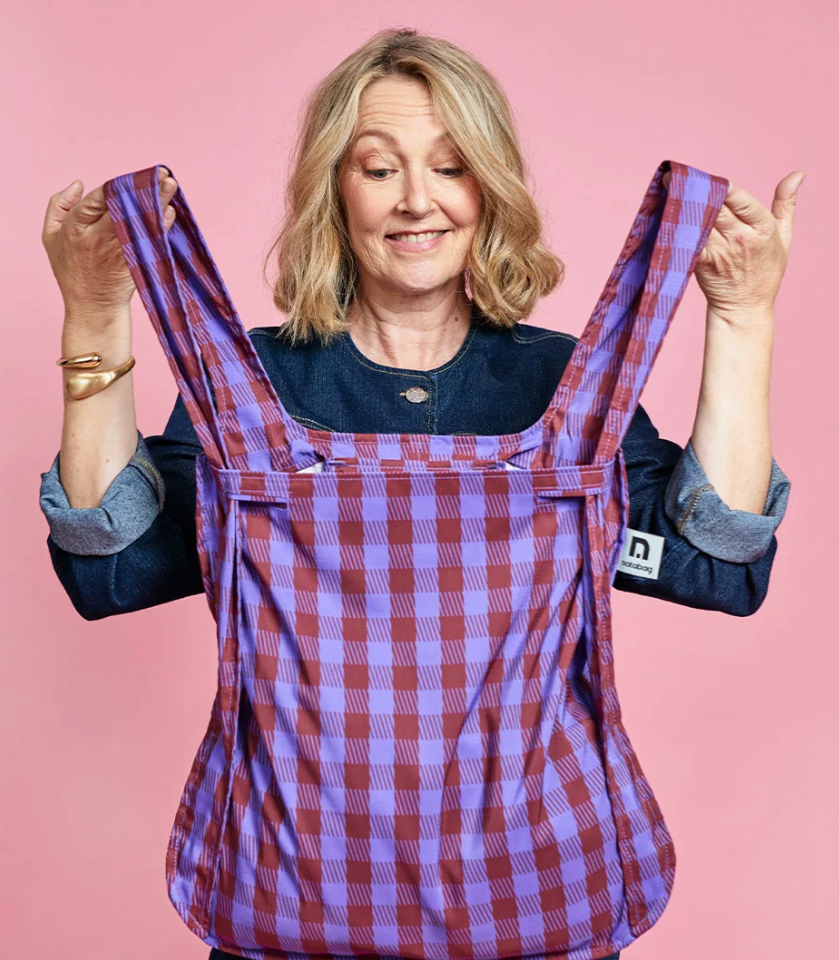 A woman holding up a baby carrier with a purple and red checkered pattern against a solid pink background. The brand tag on the side reads 'nuna'.