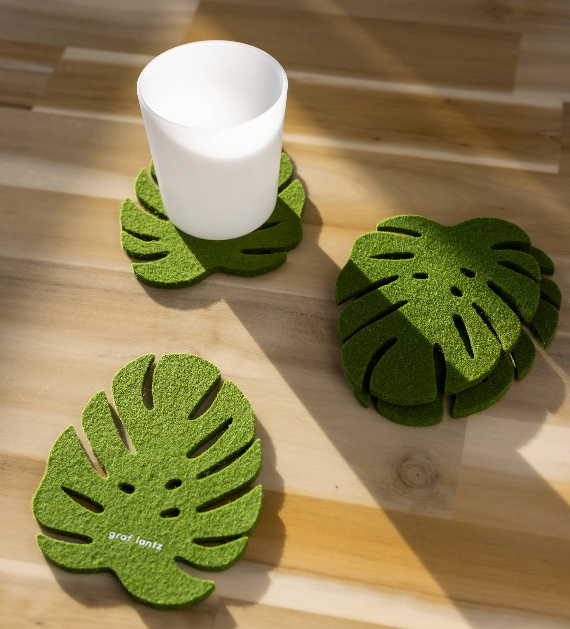 Three green monstera leaf-shaped felt coasters on a light wood surface, with one coaster holding a white cup. The word 'grofents' is printed on the front of one coaster.