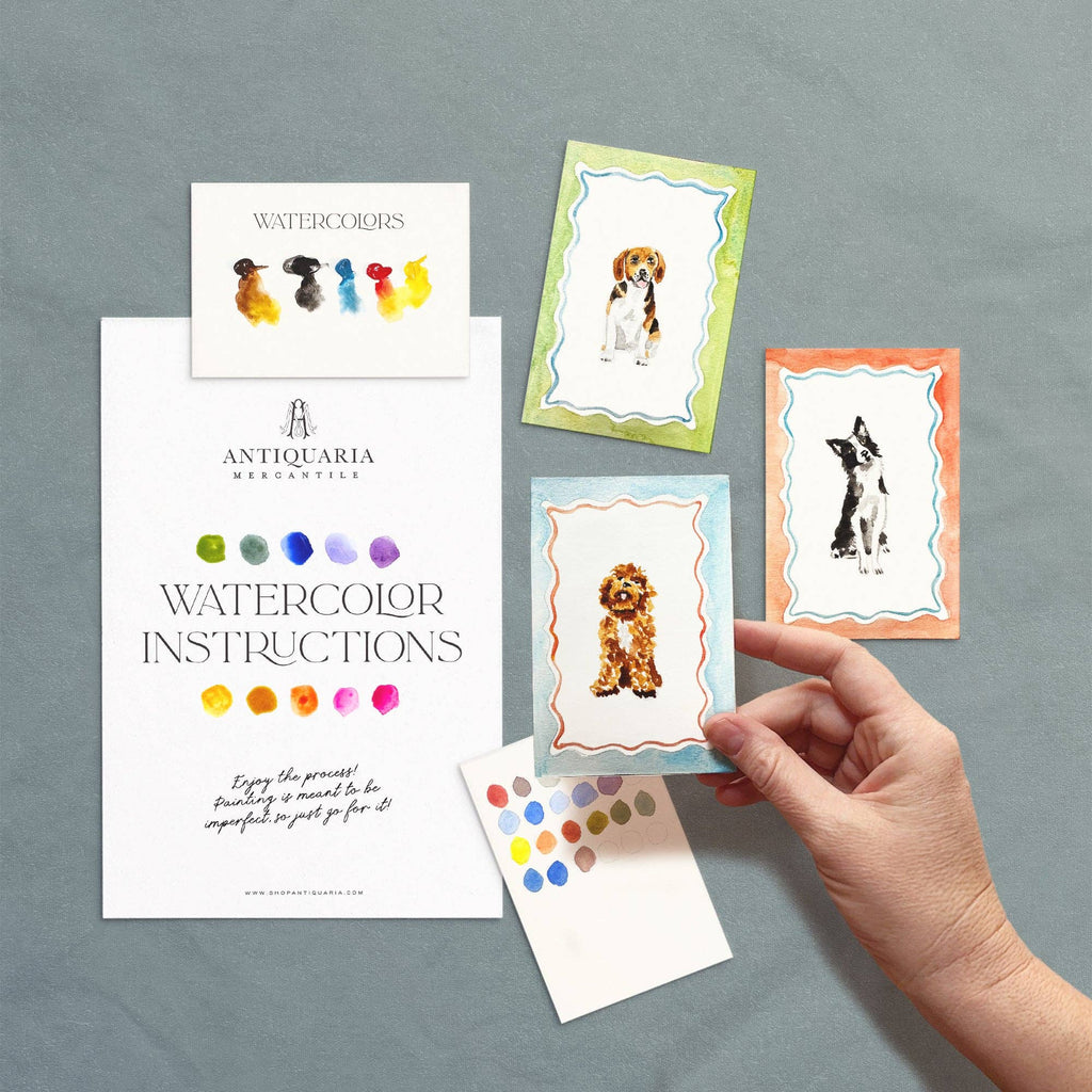 Hand holding a watercolor instruction card with dog illustrations on a gray background