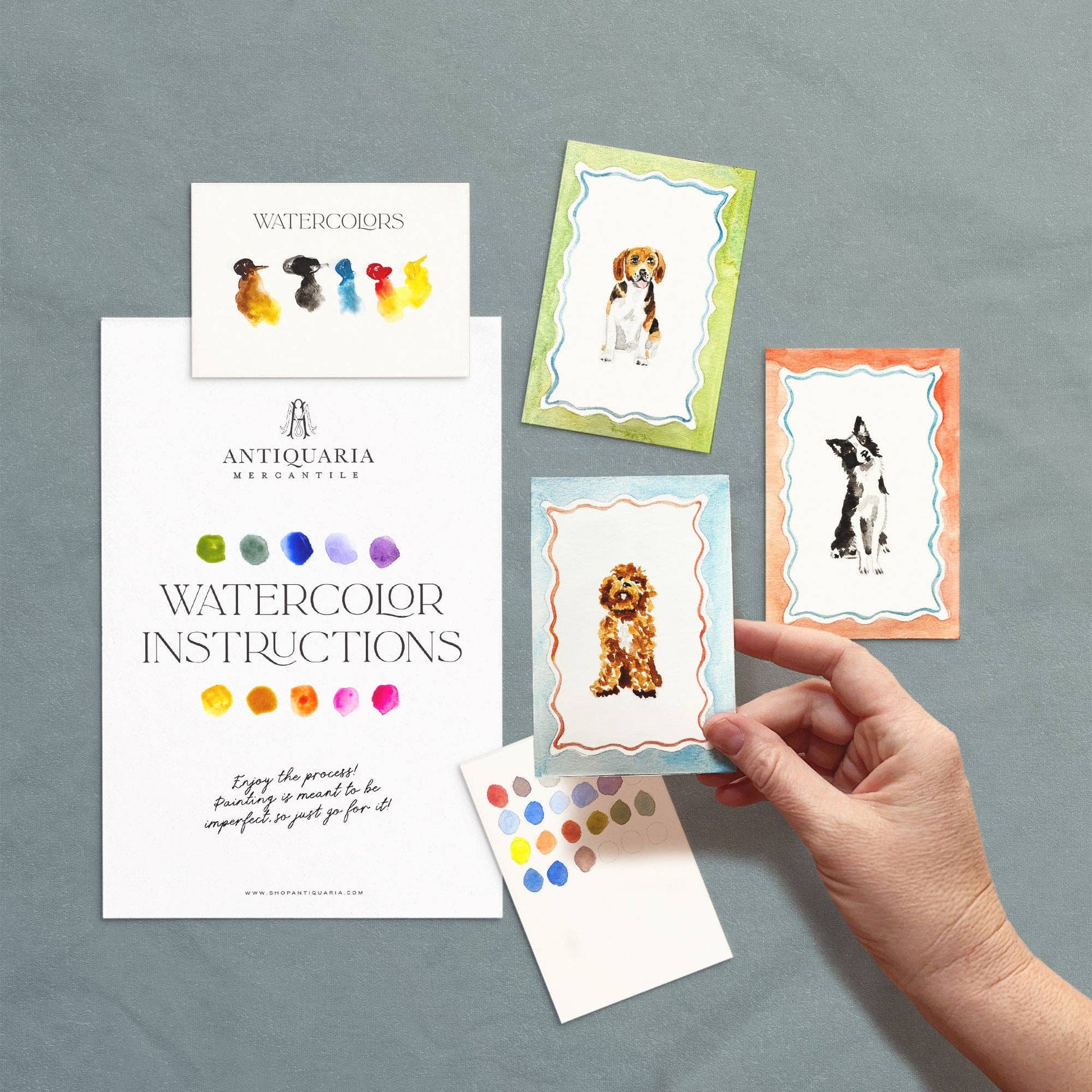 Hand holding a watercolor instruction card with dog illustrations on a gray background