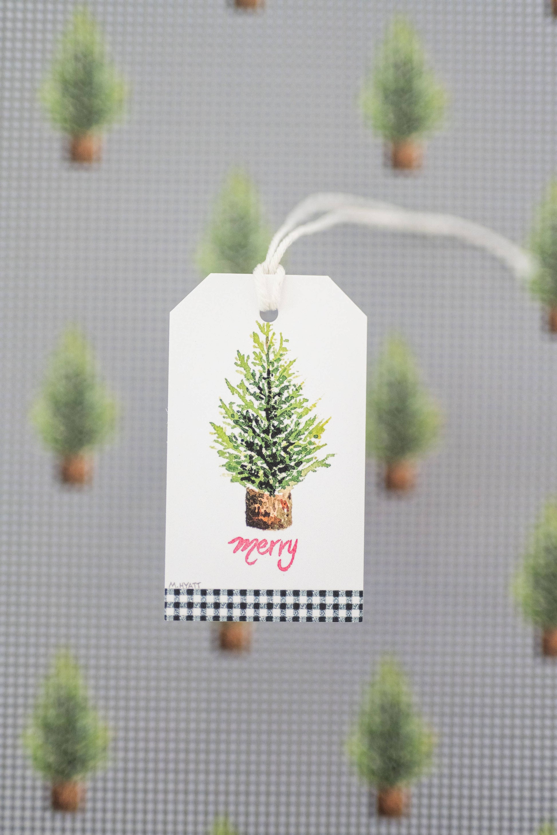 A white gift tag with a watercolor green Christmas tree and the word 'merry' in pink script, hanging against a gray patterned background of small green trees.
