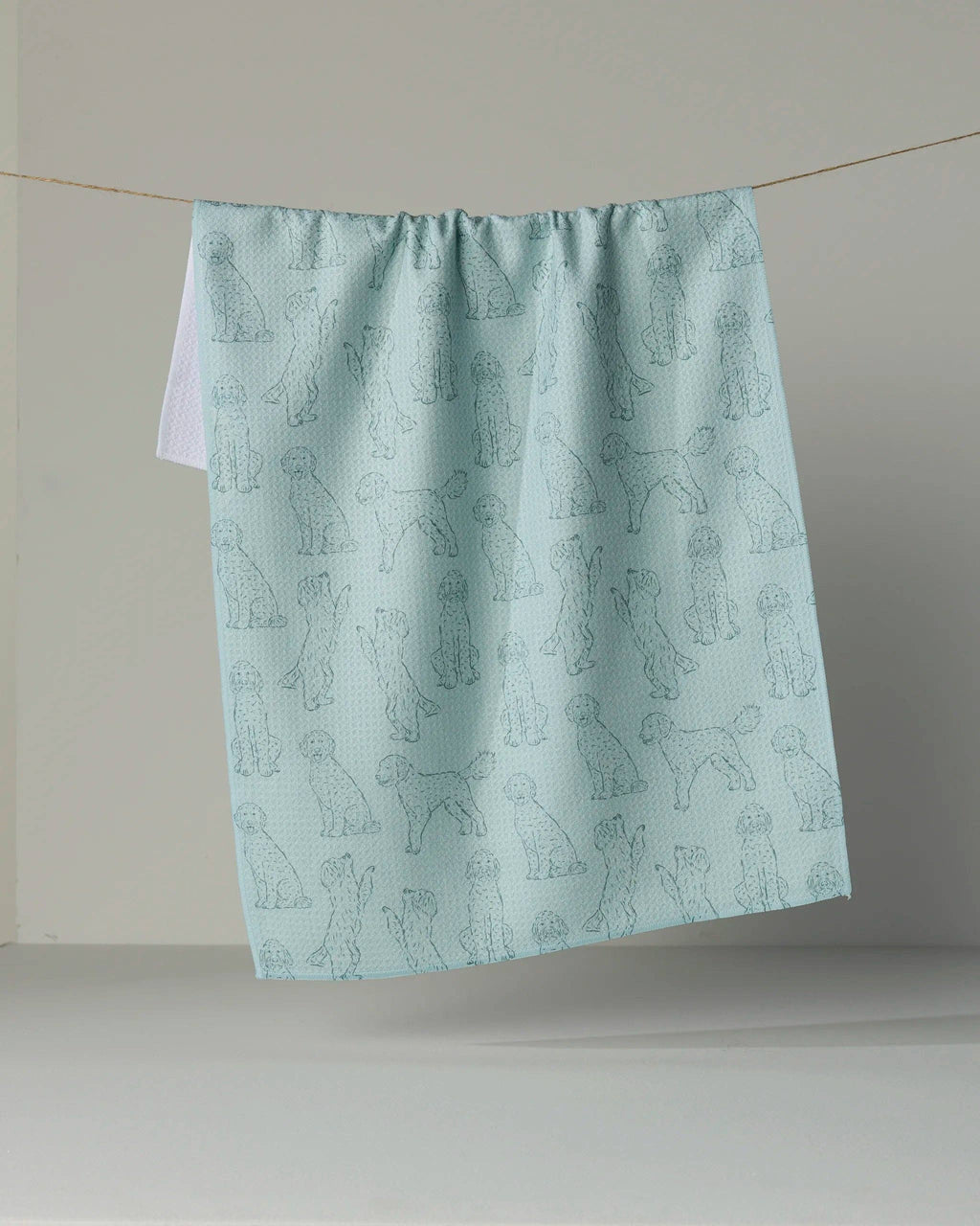 A light blue kitchen towel with a repeating pattern of dog silhouettes, hanging from a string against a plain beige wall.