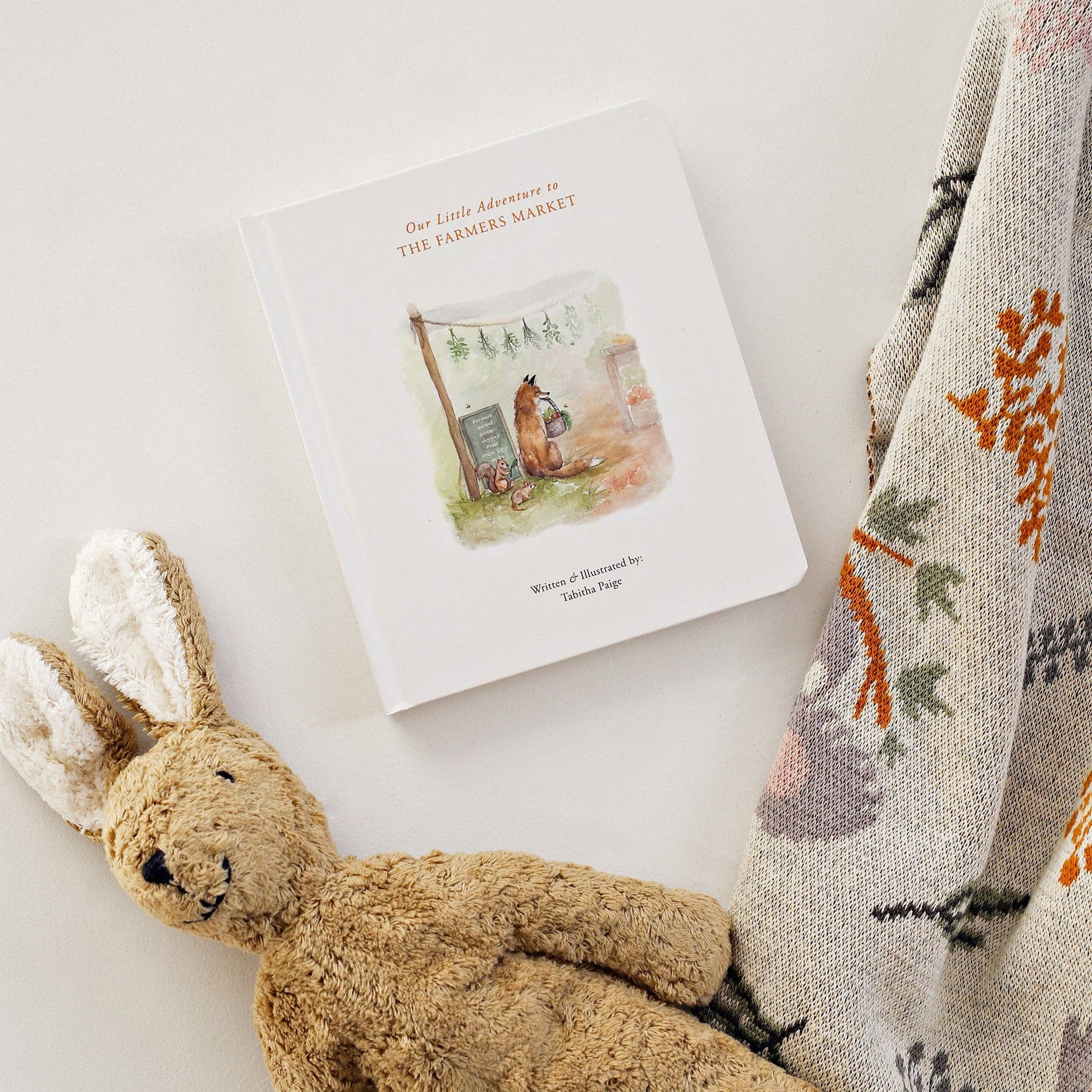 Children's book titled 'Our Little Adventure to the Farmer's Market' with a plush rabbit toy and patterned blanket on a light background.
