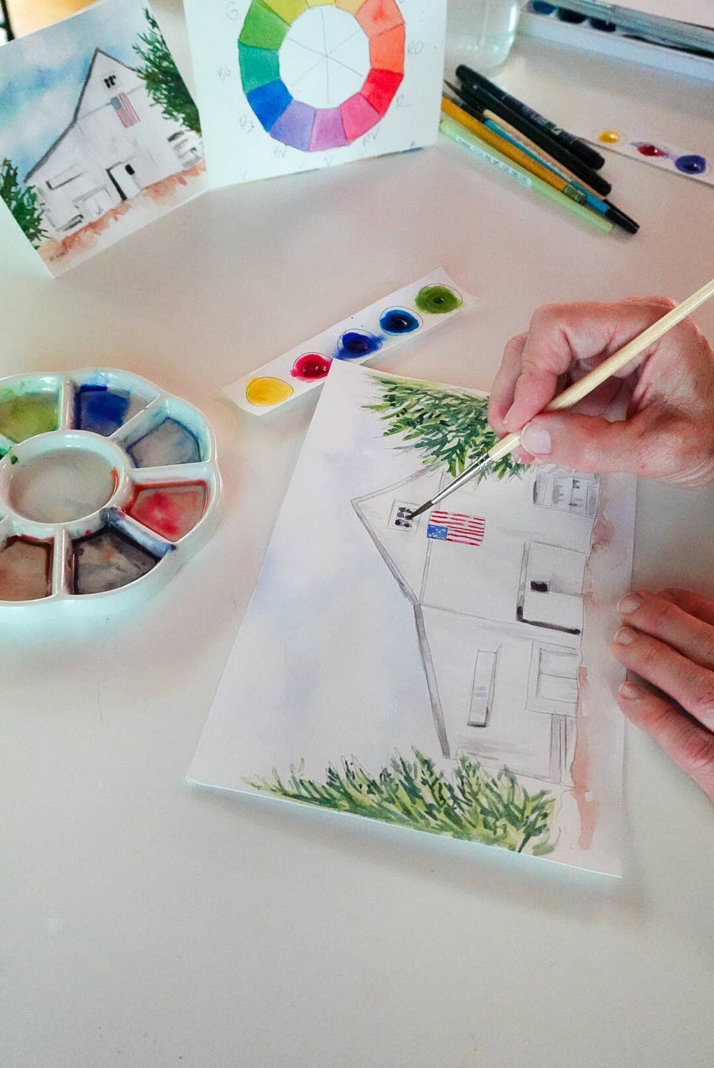A person's hand holding a paintbrush, painting a house scene on a canvas. The scene includes a watercolor palette, a color wheel card, and other art supplies on a white table.