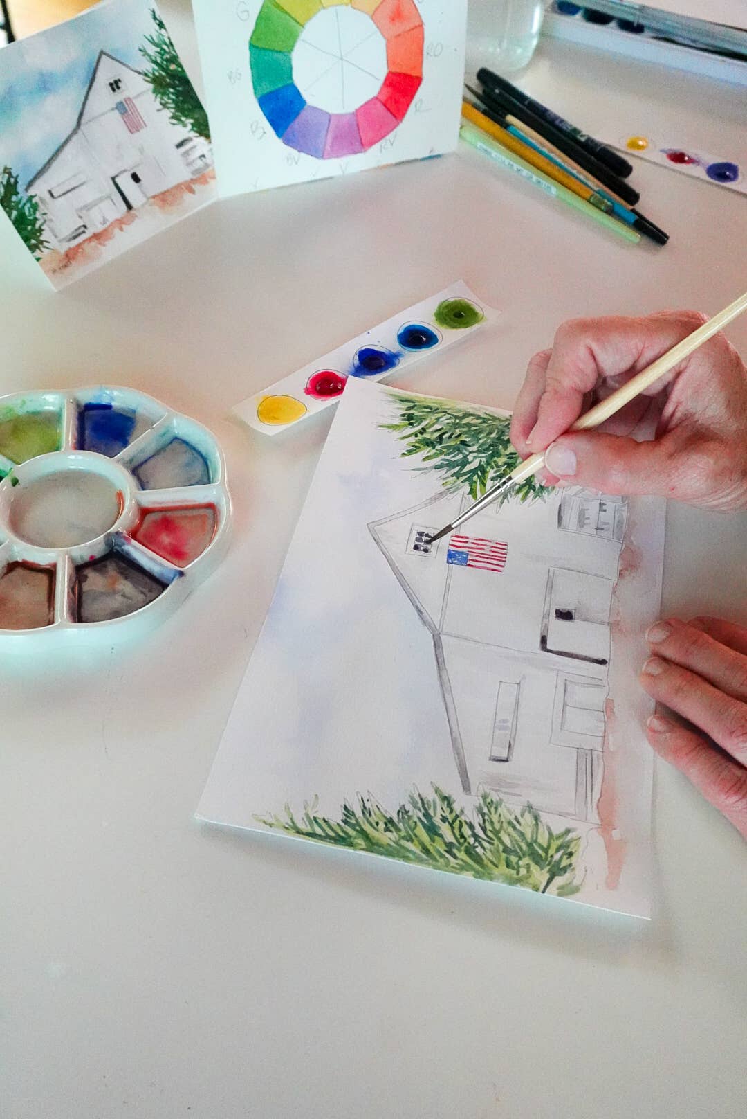 A person's hand holding a paintbrush, painting a house scene on a canvas. The scene includes a watercolor palette, a color wheel card, and other art supplies on a white table.