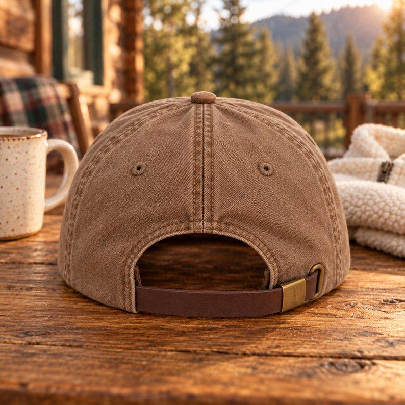 A brown cotton baseball cap shown from the back, resting on a rustic wooden table in a cozy outdoor cabin setting with a mug and blanket nearby.