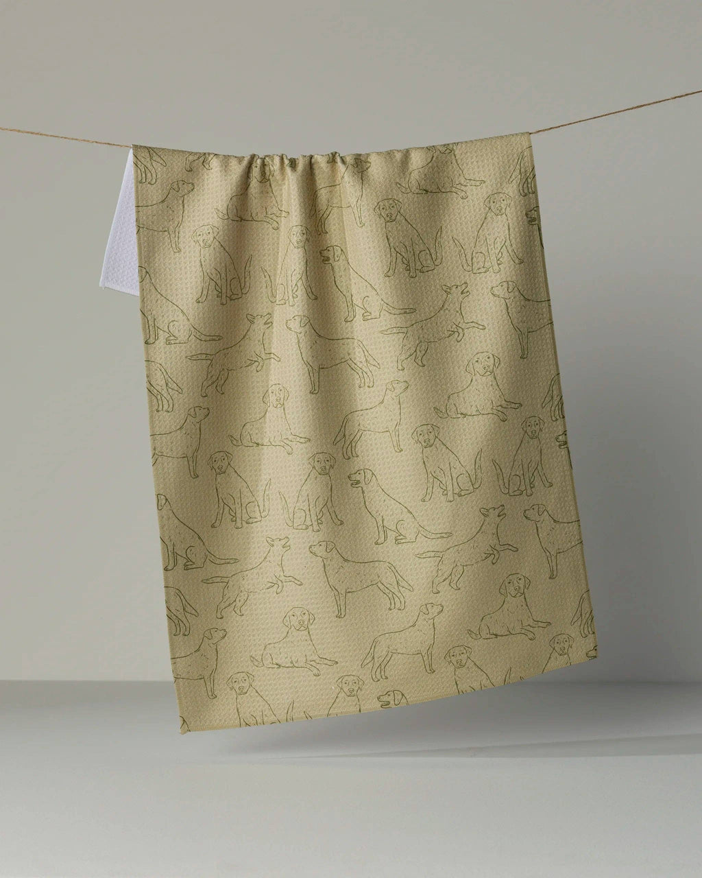 A beige kitchen towel with an all-over print of dog line drawings, hanging from a string against a neutral gray background.