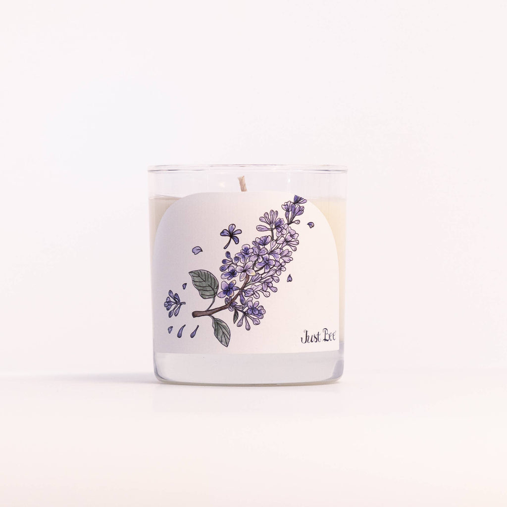 A clear glass jar candle with white wax and a white label featuring a purple floral illustration and the text 'Just Luv'.