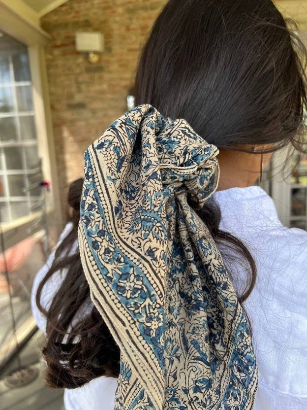 A dark brown hair accessory with a blue and beige floral block-print pattern, tied around a person's dark hair in a porch setting.