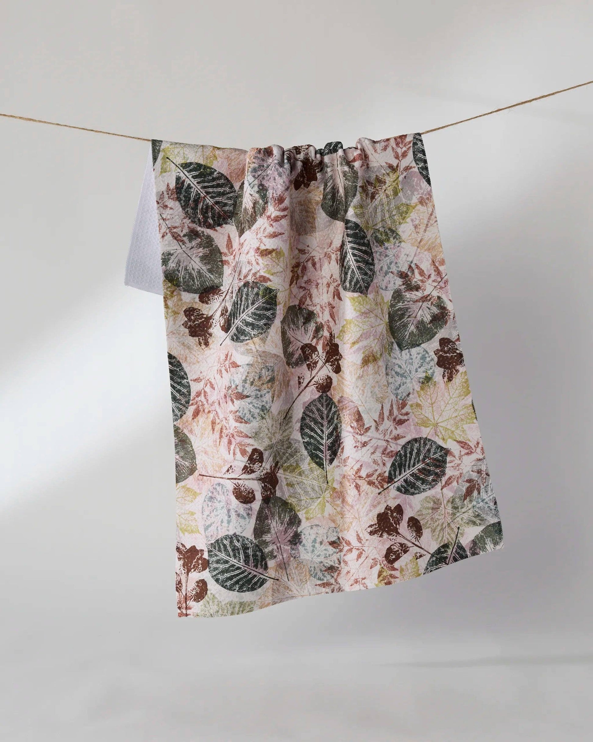 A lightweight fabric curtain panel with a botanical leaf pattern in muted green, brown, and dusty pink on a cream background, hanging from a thin wire against a plain light gray studio backdrop.