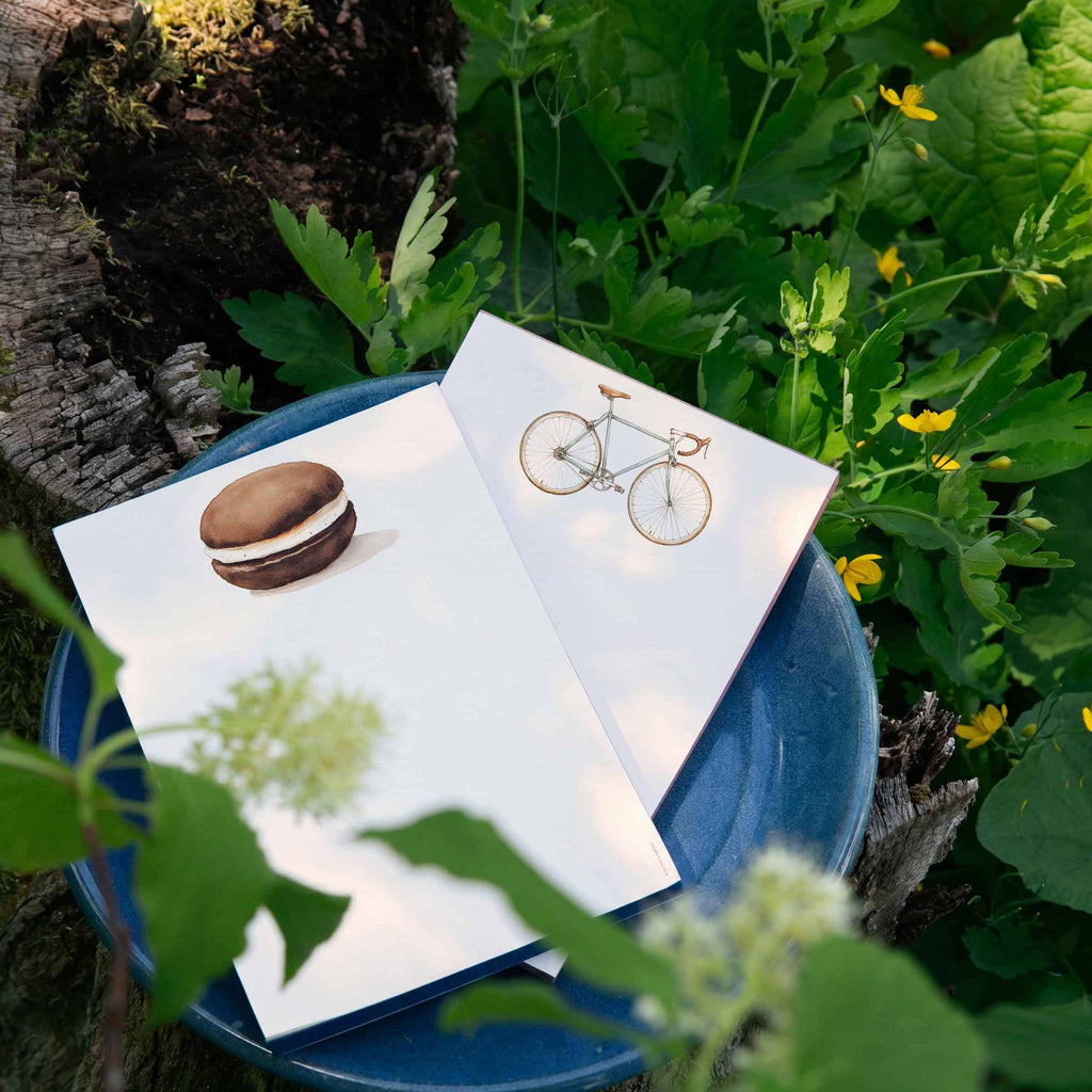 Two greeting cards with macaron and bicycle design on a blue dish surrounded by greenery.