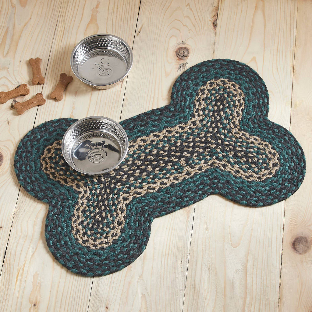 A braided bone-shaped rug in dark teal and beige sits on a light wood plank floor, with two stainless steel dog bowls placed on top and a few bone-shaped props nearby.