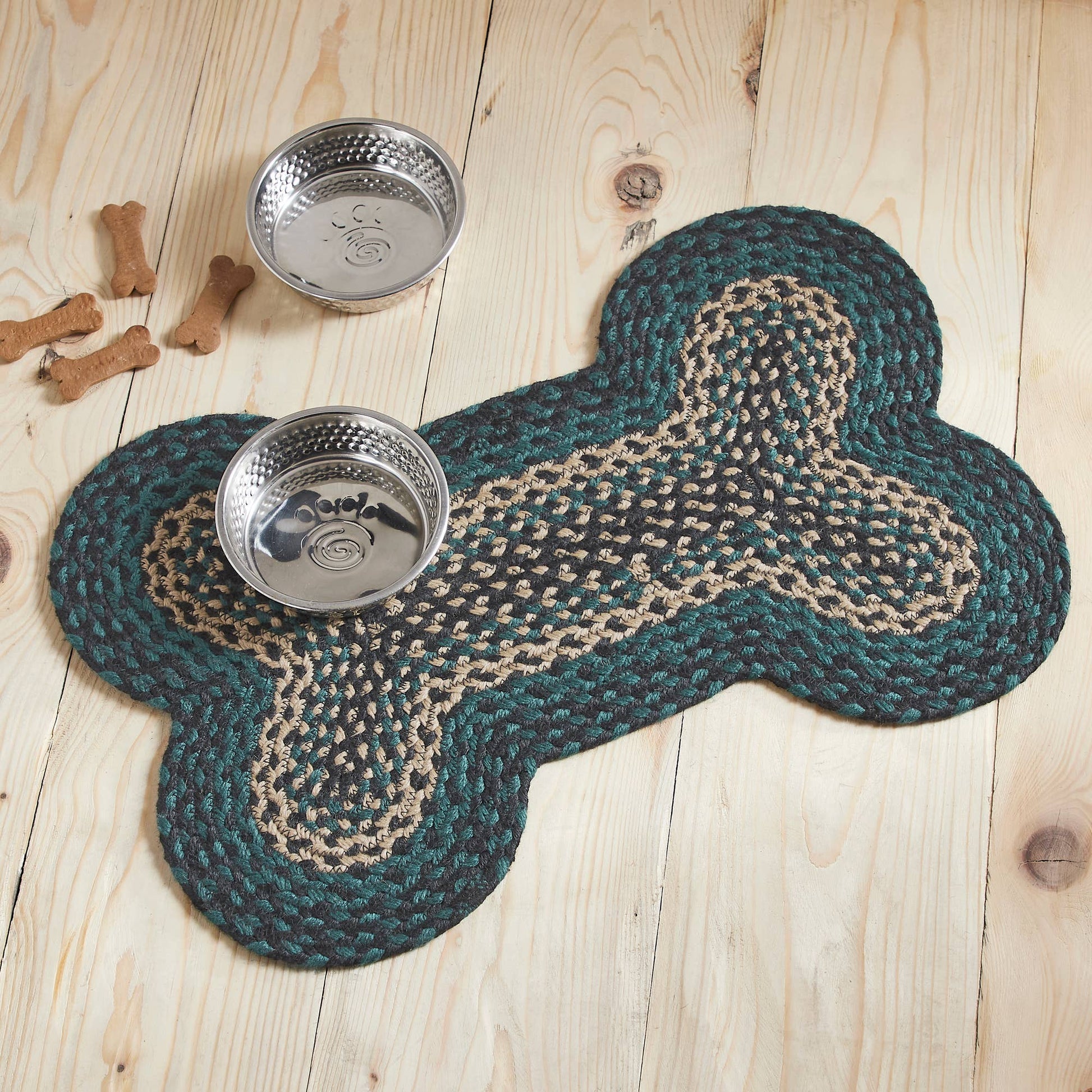 A braided bone-shaped rug in dark teal and beige sits on a light wood plank floor, with two stainless steel dog bowls placed on top and a few bone-shaped props nearby.
