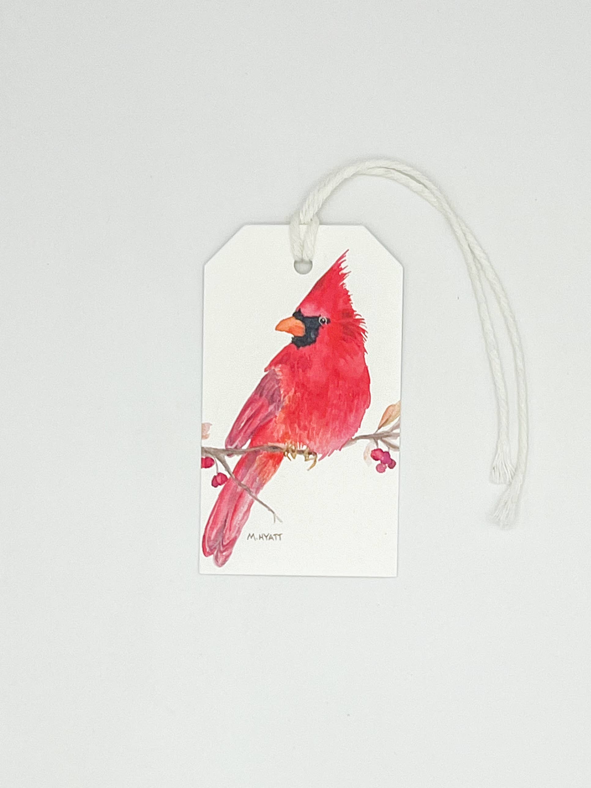 A white gift tag with a hanging white string, printed with a watercolor-style red cardinal perched on a branch with small red berries. The tag is photographed against a plain light gray background.
