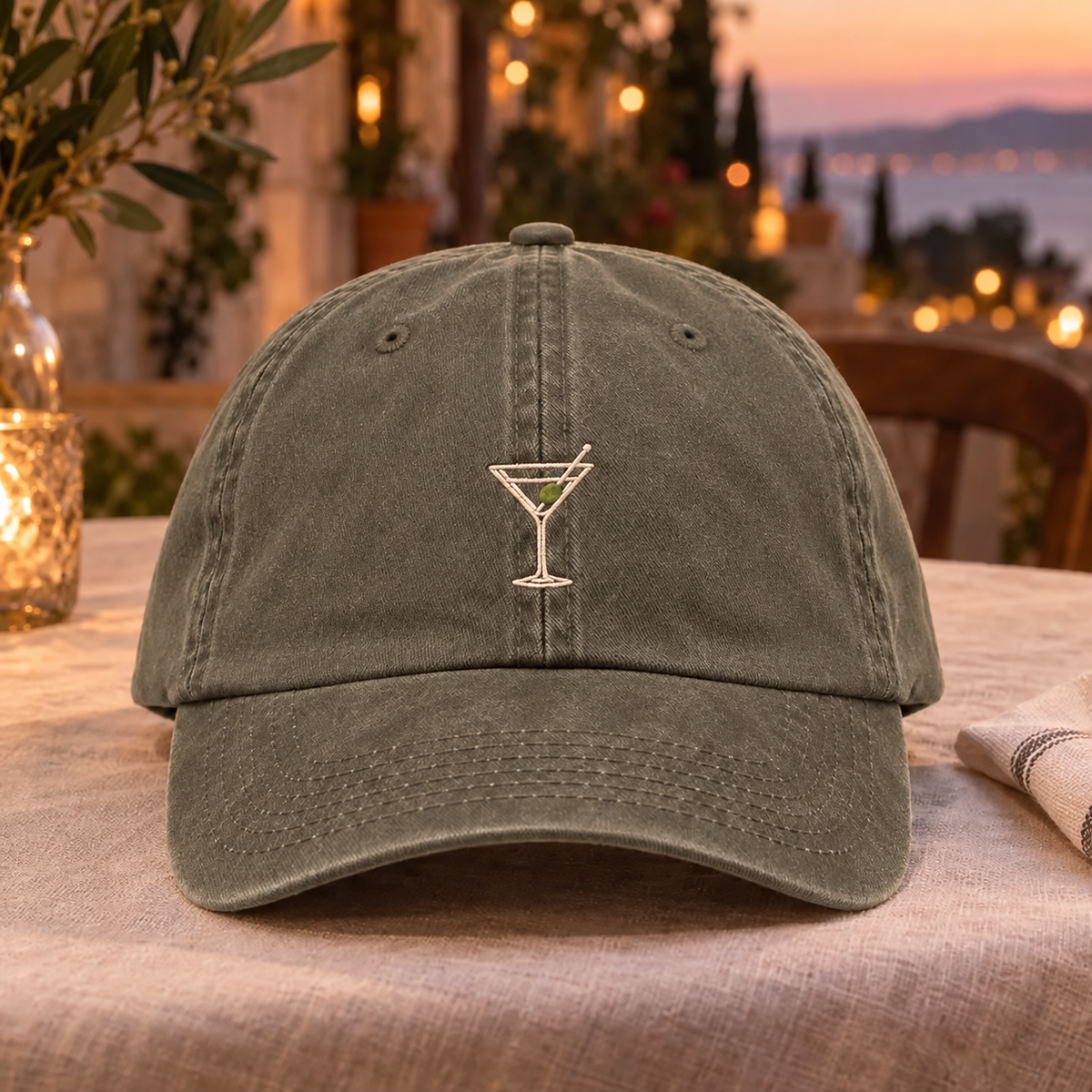 An olive green washed cotton baseball cap with an embroidered martini glass on the front, displayed on a table in a warm, softly blurred outdoor setting.