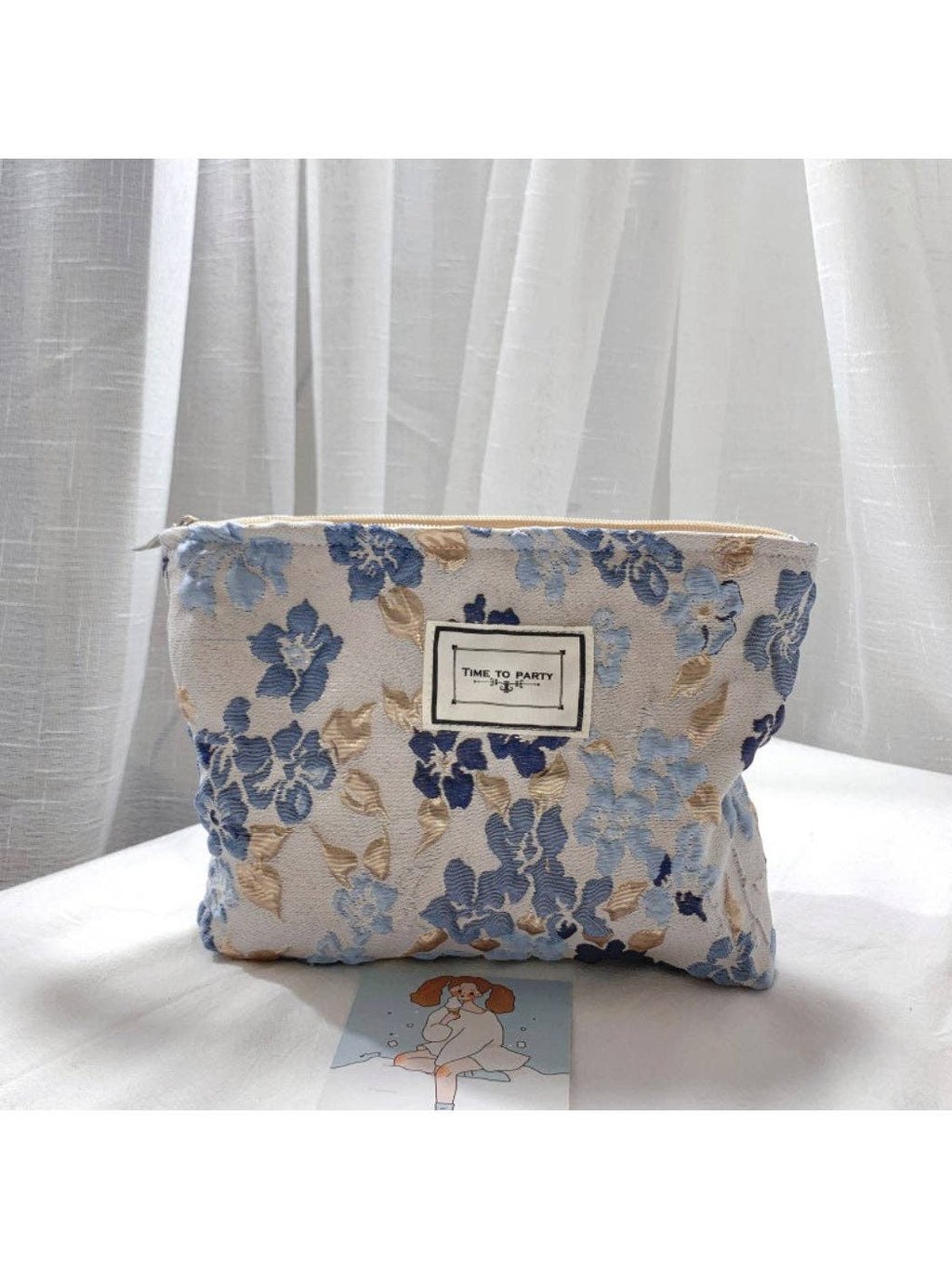 A fabric cosmetic pouch with a blue and beige floral pattern, featuring a central white label that reads 'time to travel', set on a white surface in front of sheer white curtains.