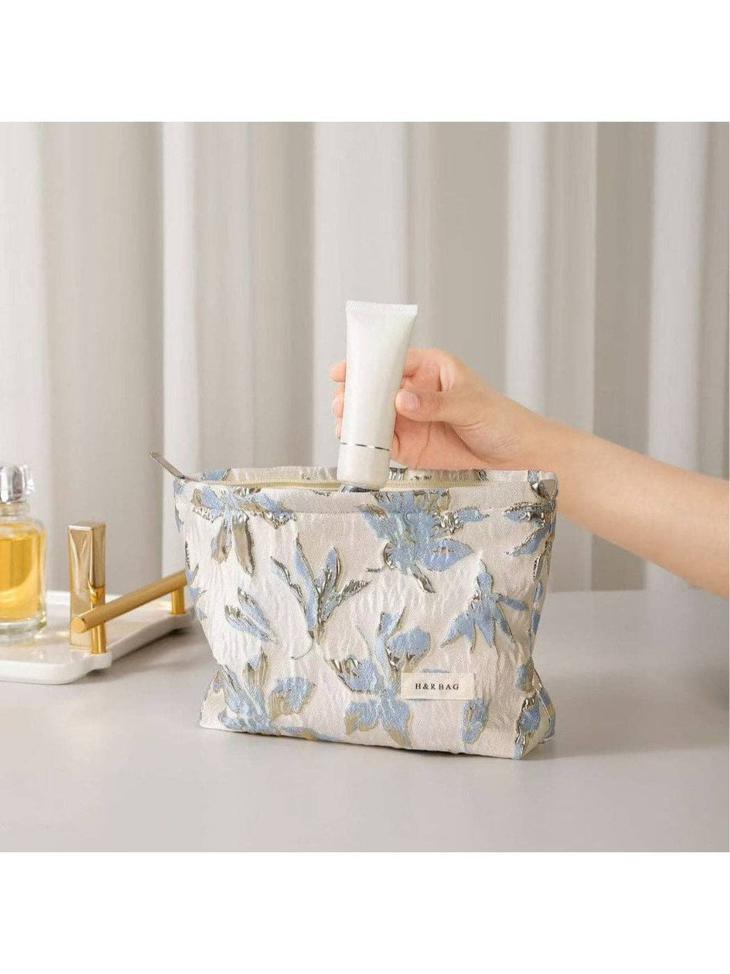 Hand holding a tube of cream above a floral-patterned pouch on a neutral background