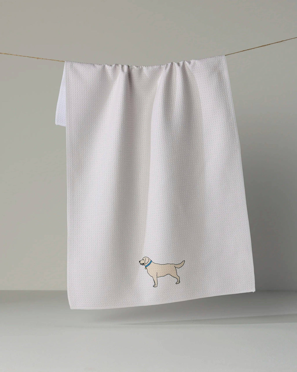 A white waffle-weave kitchen towel hanging from a thin string against a neutral gray background, featuring a small embroidered cartoon dog wearing a blue collar near the bottom.