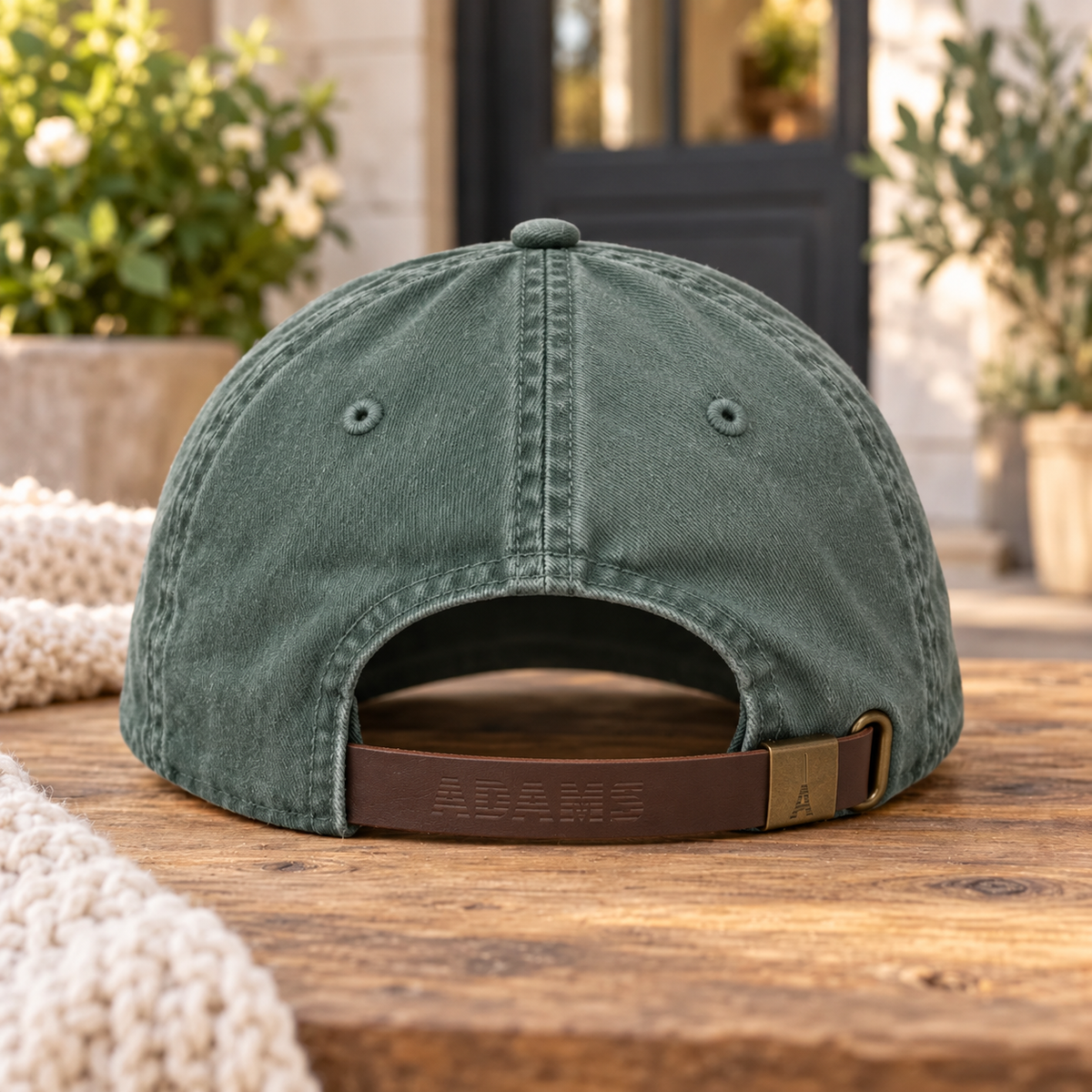 A washed dark green baseball cap shown from the back, resting on a rustic wooden table with a brown leather strap closure. The background features a softly blurred outdoor patio setting with potted plants and a doorway.