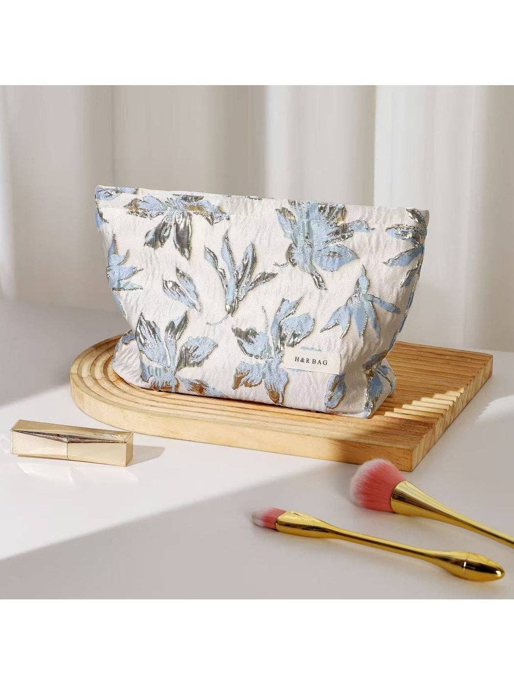 Floral-patterned makeup bag on a wooden tray with lip gloss and brushes on a white surface.