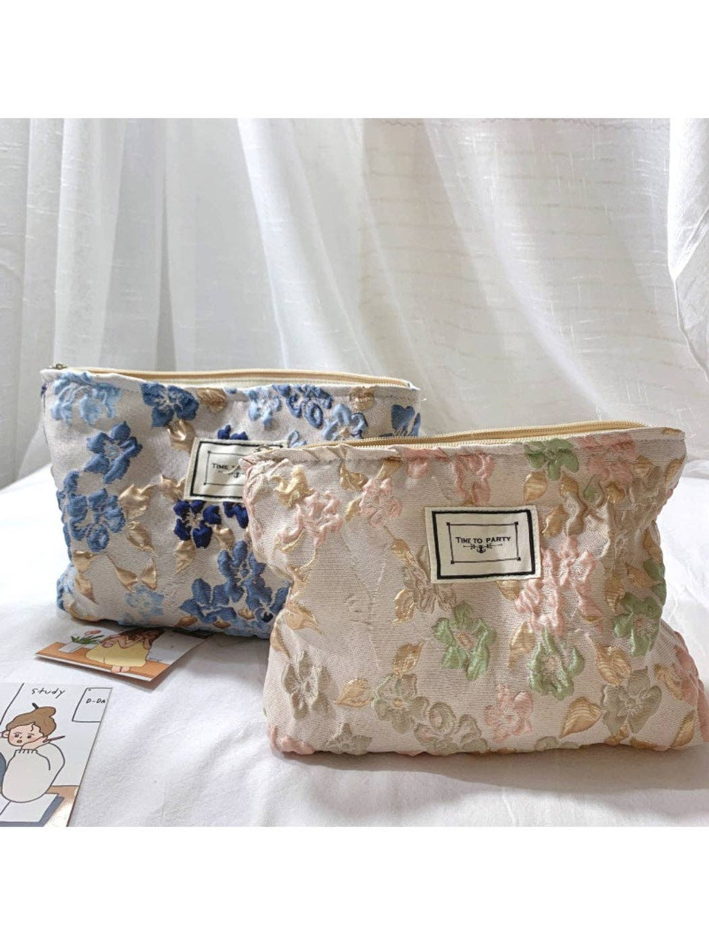 Two floral embroidered cosmetic pouches with gold-tone zippers, displayed on a white fabric surface in front of sheer white curtains. One pouch is light blue with blue floral embroidery, and the other is cream with pink and green floral embroidery.
